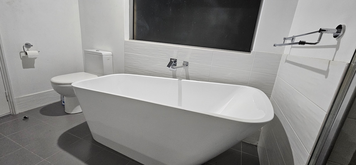 A clean and modern bathroom is displayed, featuring a freestanding bathtub alongside a compact toilet. Neutral-toned tiles cover the floor and walls, while a large window allows for natural light to enter the space.
