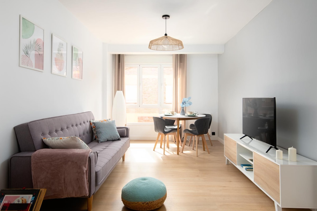 A bright living room features a modern design with a grey sofa, a round dining table with four chairs, and a large window allowing natural light. Minimalist artwork decorates the walls, and a television is positioned on a white media console.