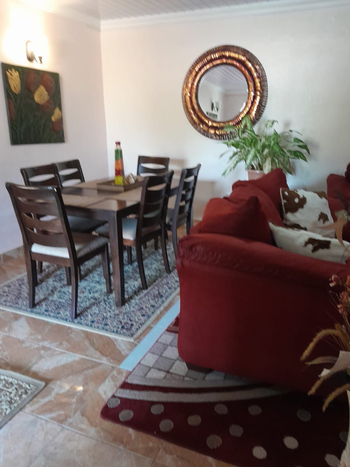 A dining area is showcased with a wooden table surrounded by six chairs, positioned atop an area rug. A circular mirror adorns the wall, with a decorative plant adding a touch of greenery, and a cozy seating area features a red sofa with patterned cushions.