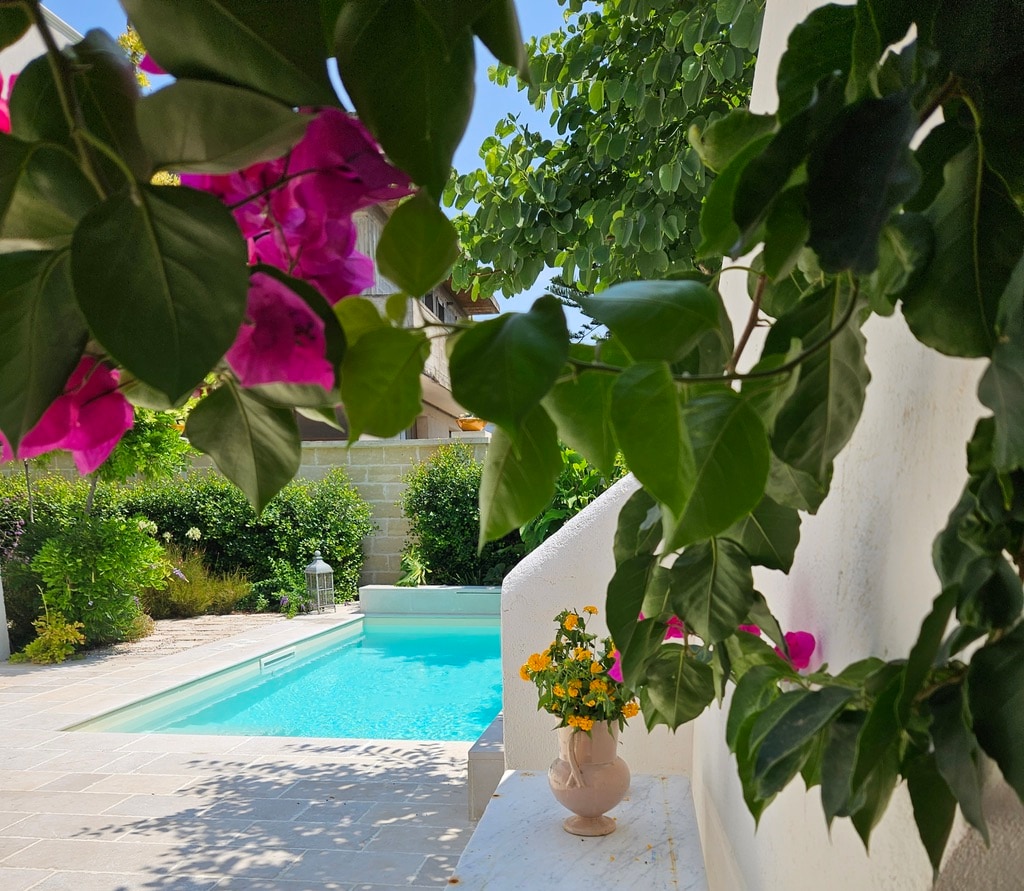 A serene garden setting is visible, featuring a private saltwater pool surrounded by lush greenery and blooming flowers. Colorful potted plants are positioned near the pool, enhancing the tranquil atmosphere in the outdoor space.