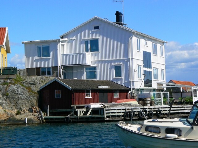 A two-story white house with large windows overlooks a rocky shoreline. A red wooden building is visible at the water's edge, complemented by a small boat docked nearby. The scene captures the coastal setting, with clear blue skies and water reflecting the surroundings.