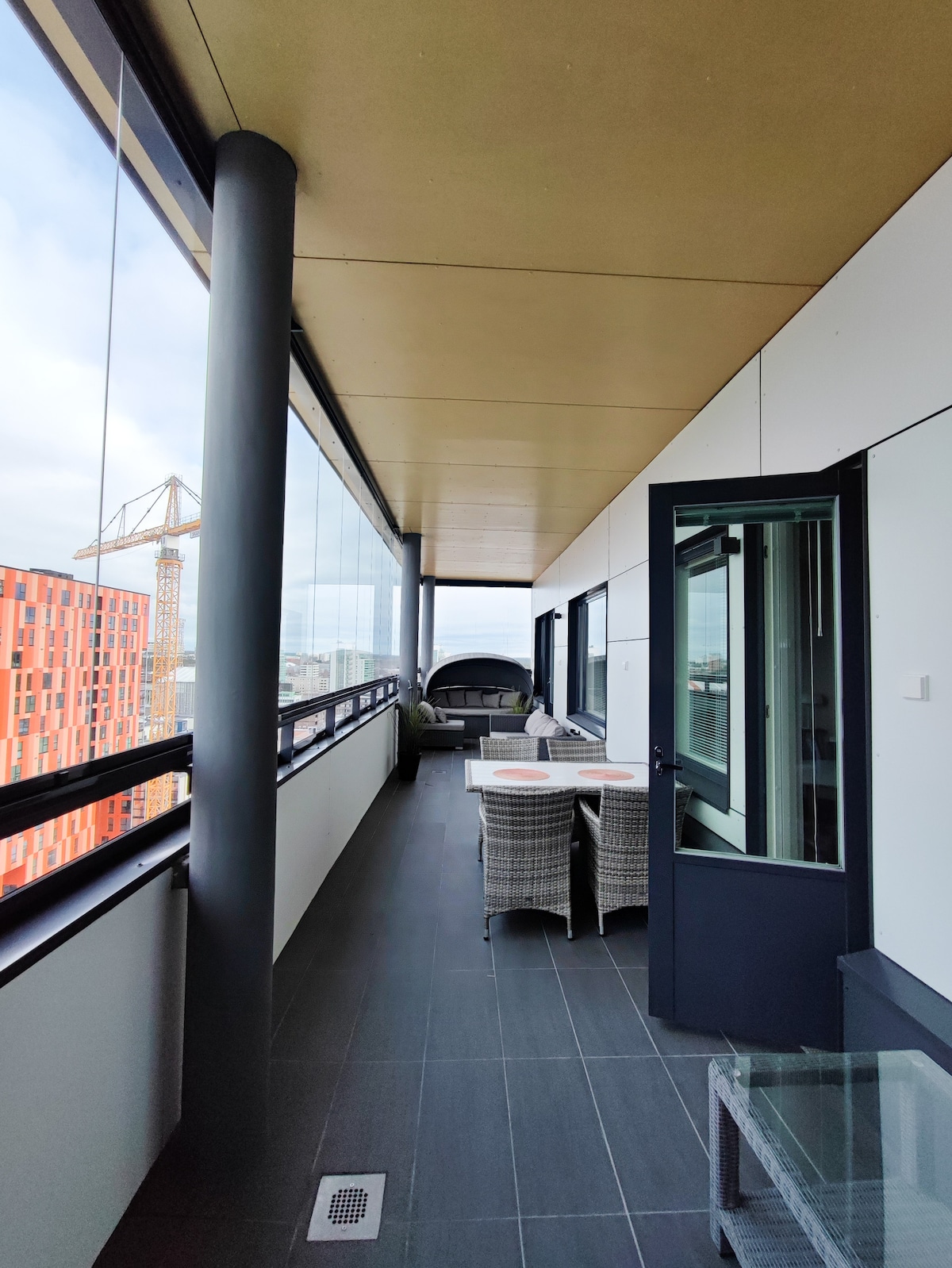 A spacious glass-enclosed balcony features modern outdoor furniture, including a dining table with matching chairs. Large glass panels allow ample natural light and city views, while an adjacent door provides access to the residing unit. The flooring is sleek, enhancing the contemporary design.