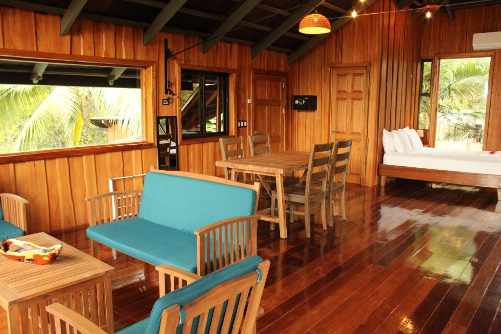 A spacious interior features wooden walls and floors, complemented by large windows providing views of the surrounding nature. A seating area includes turquoise chairs and a wooden table, while a cozy sleeping nook is visible in the background, highlighted by natural light.