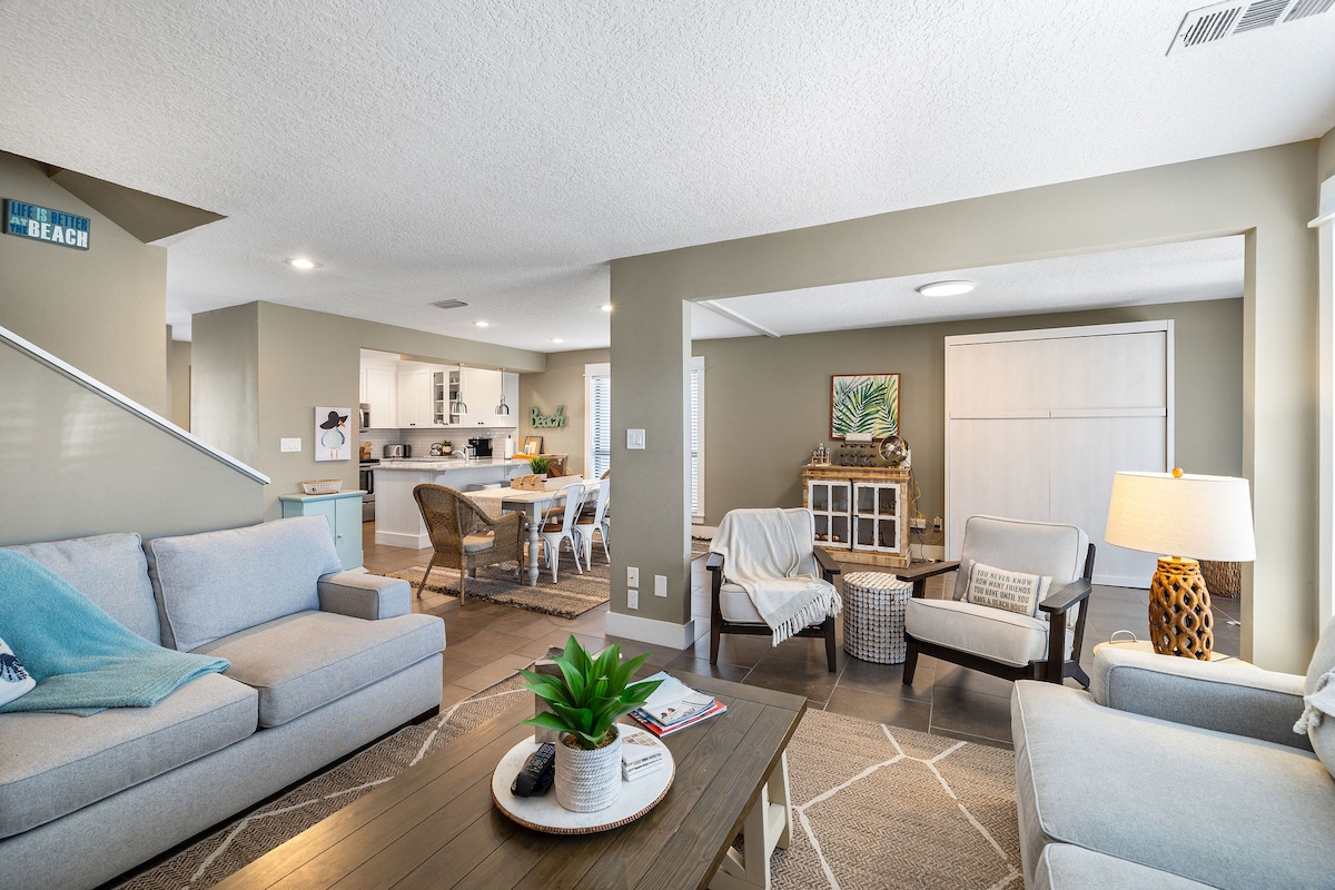 A bright living area features comfortable seating, including a spacious sofa and stylish armchairs. A wooden coffee table rests in the center, adorned with a plant. In the background, an open dining area and kitchen are visible, enhancing the spacious feel of the home.