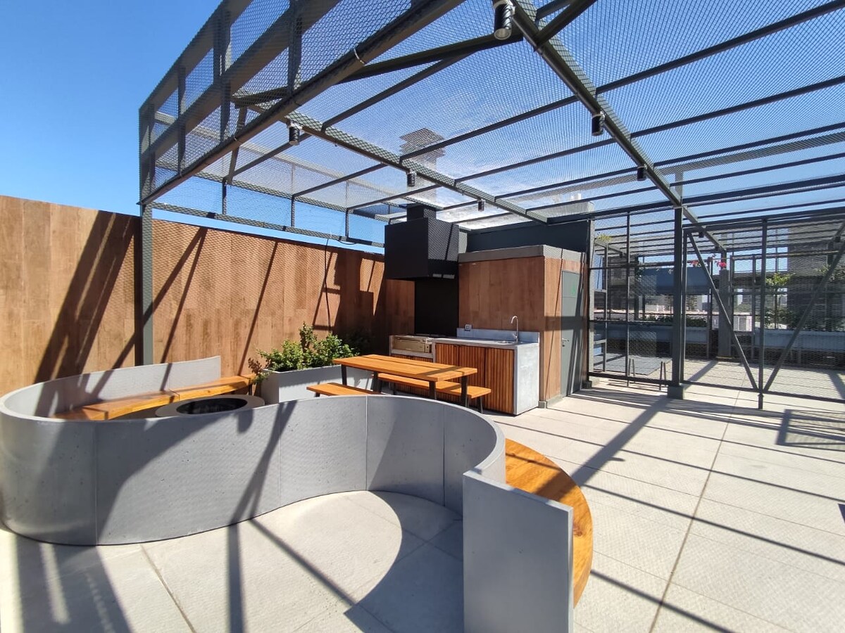 An outdoor communal area is presented featuring a circular concrete seating arrangement and wooden benches around a central fire pit. A barbecue grill and countertop are located under a protective roof, surrounded by sunlit open space with wooden paneling and greenery.