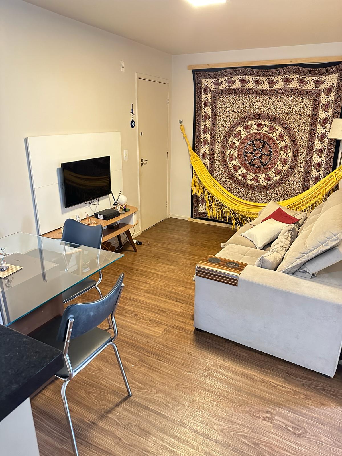 A living area features a light-colored sofa positioned beside a yellow hammock, creating a cozy setting. A modern TV unit is mounted on the wall, accompanied by a small desk with a chair. A decorative tapestry adds a touch of character to the room.