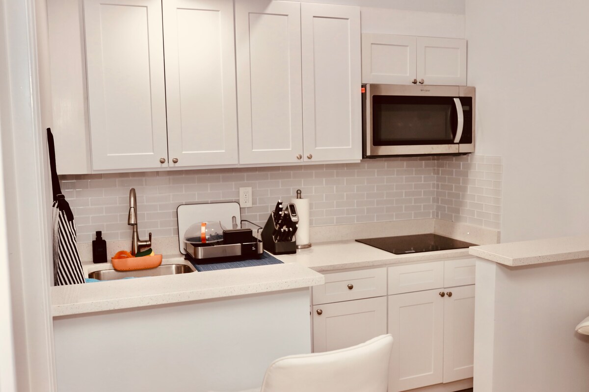 A modern kitchen area is presented with light cabinetry and a smooth countertop. Stainless steel appliances, including a microwave and stovetop, are visible. A sink is positioned under a window, and a small decorative item adds a touch of detail to the countertop.