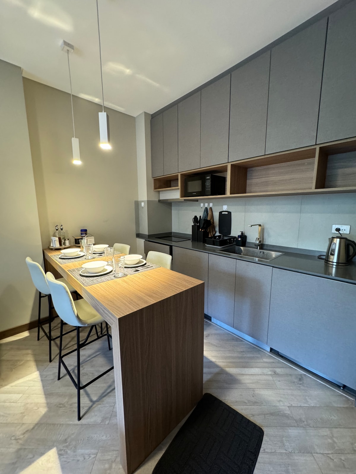 A modern kitchen area features a wooden breakfast bar with four stools, set with plates and glassware. Sleek cabinetry above and below provides ample storage, while a small kitchen appliance area includes a kettle and microwave. Soft lighting from two hanging fixtures enhances the space.