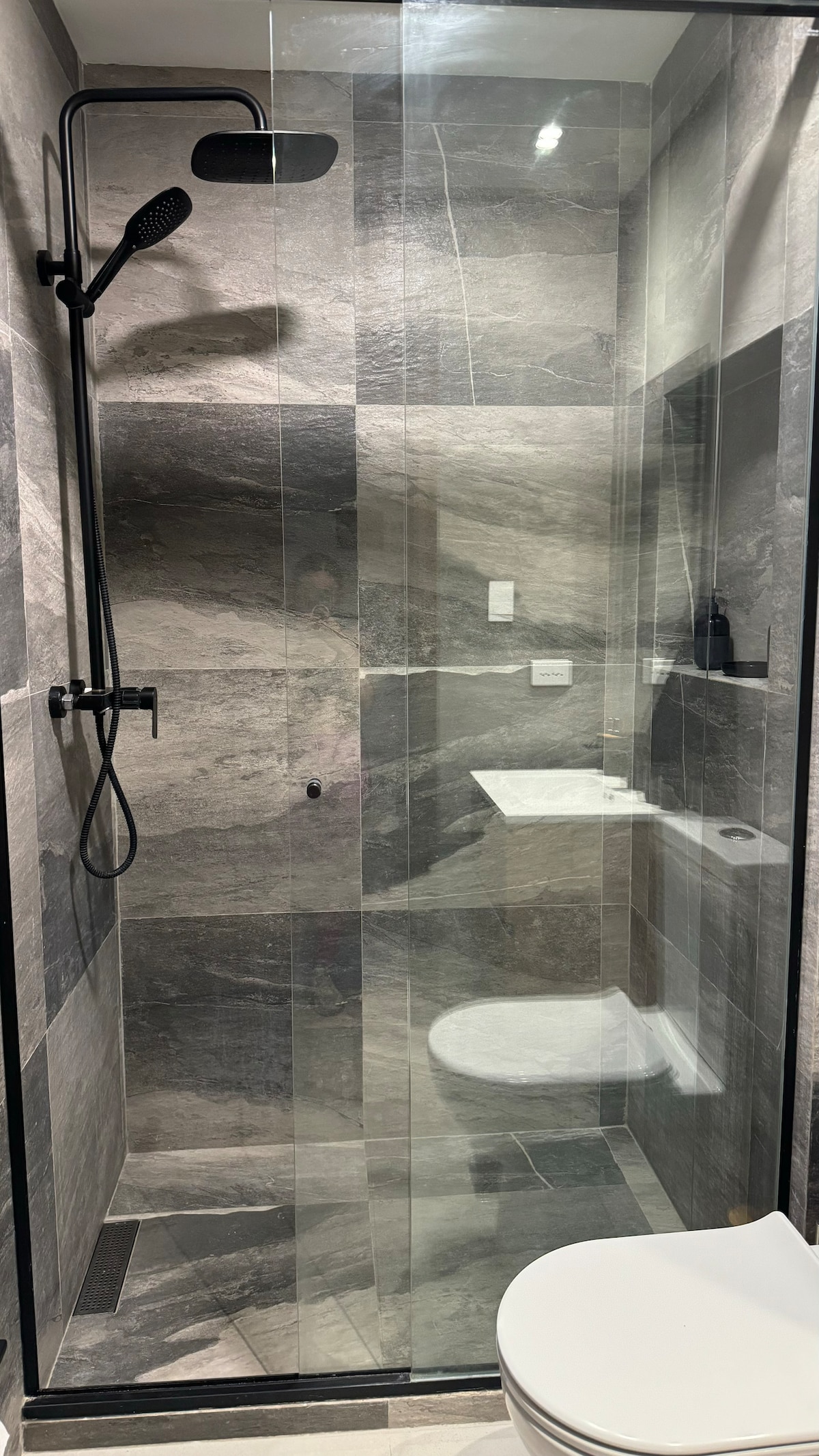 A sleek bathroom shower area features a glass enclosure and elegant grey stone tiles. A rainfall showerhead is positioned above, with a built-in bench for convenience. The minimalist design promotes a clean and modern aesthetic, complemented by a discreet toilet positioned nearby.
