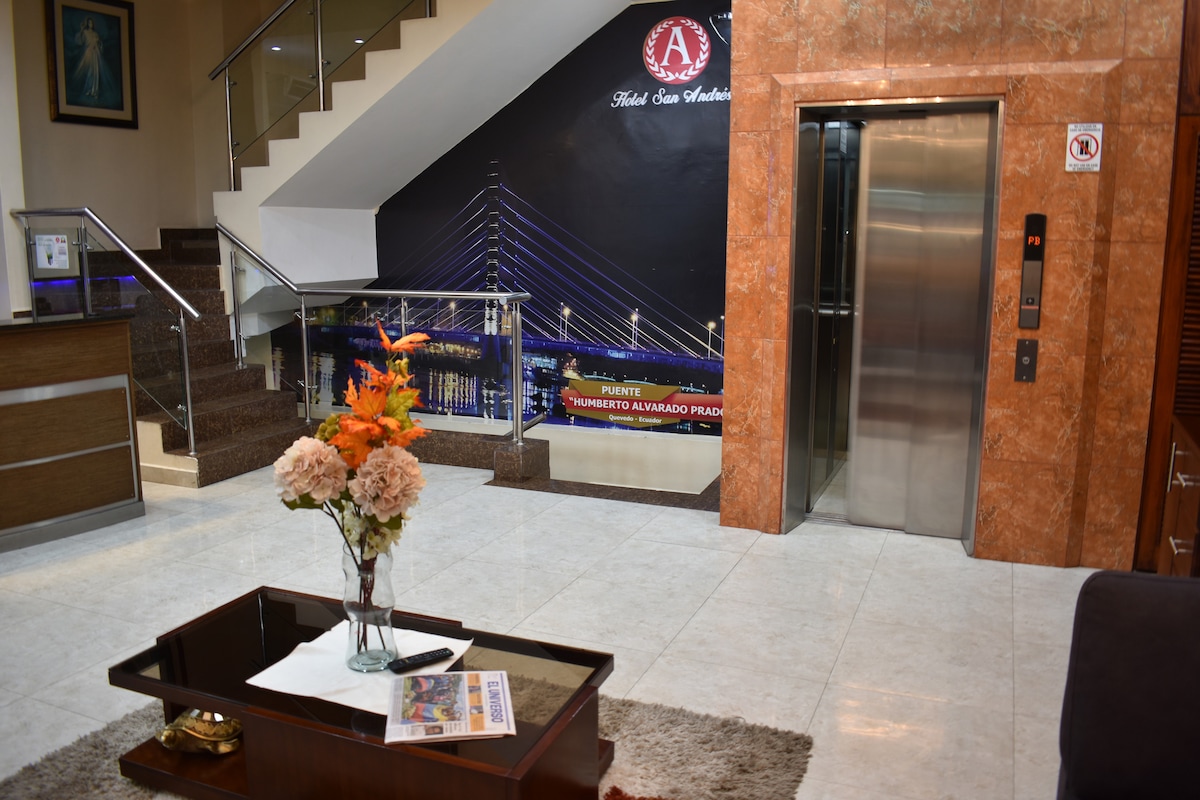 The lobby area features a sleek design with polished marble floors and a decorative table holding a vase of flowers. An elevator is visible alongside a staircase, with artwork displayed on the wall depicting a modern bridge.