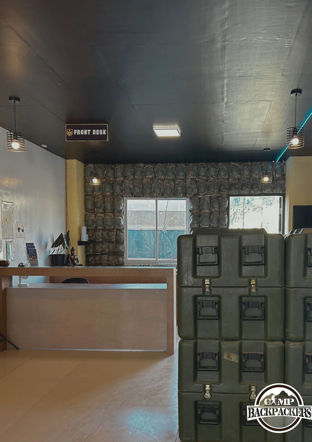 A reception area is seen featuring a front desk made of smooth wood. Behind the desk, a large window allows natural light to enter, framed by patterned curtains. Stacked green storage containers are positioned in the foreground, contributing to a functional and organized space.