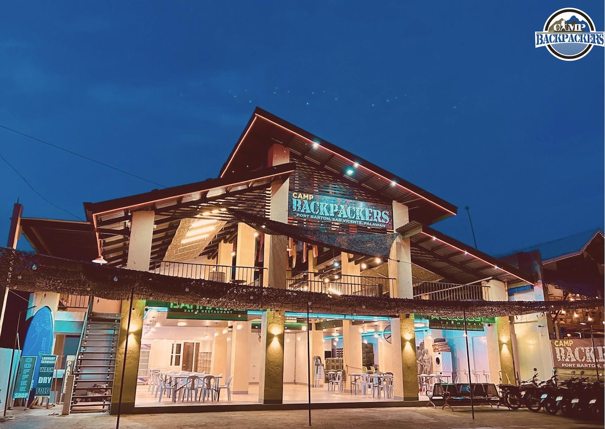 The exterior of the Camp Backpackers building is presented at dusk, showcasing a two-story structure with a spacious balcony. Outdoor seating is visible on the ground level, illuminated by soft lighting, while a clear sky fills the backdrop above the welcoming entrance.