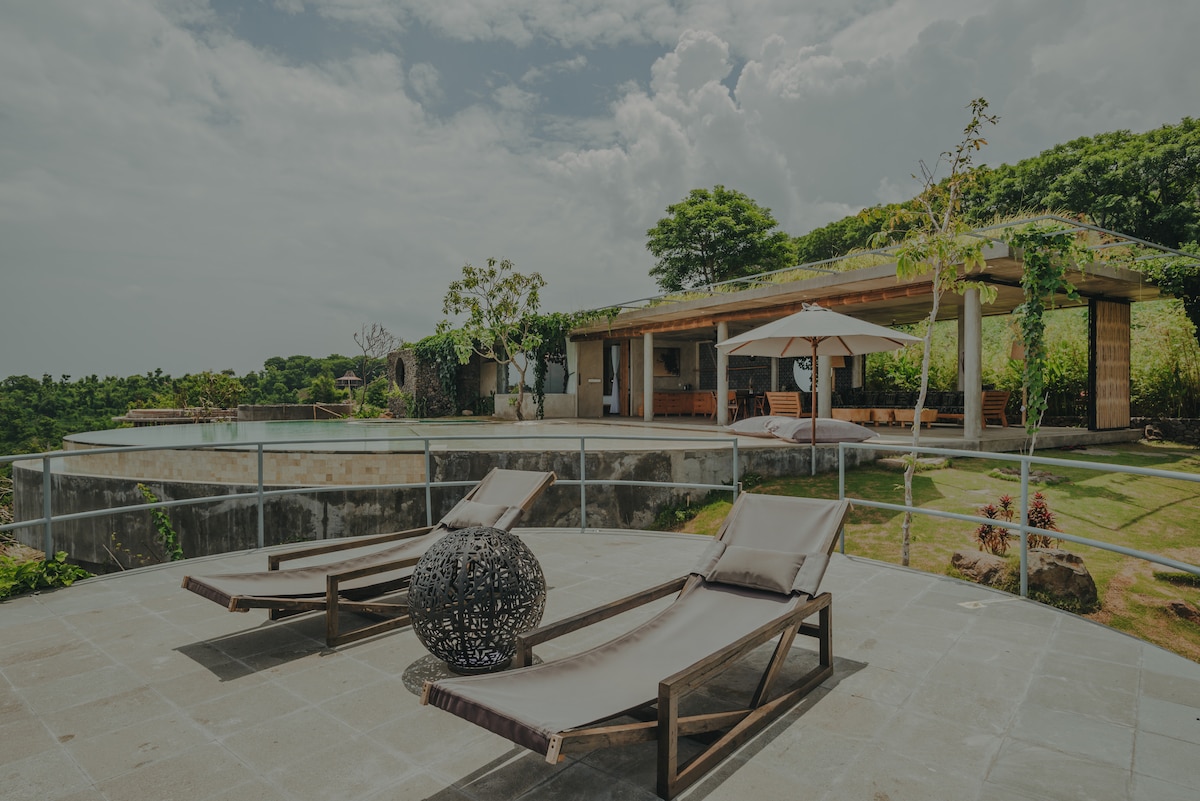 Two sun loungers are positioned on a spacious patio, facing a serene landscape. A naturally designed structure with lush greenery serves as a backdrop, featuring a large outdoor space with an umbrella and dining area nearby. The scene is complemented by a tranquil pool visible in the distance.