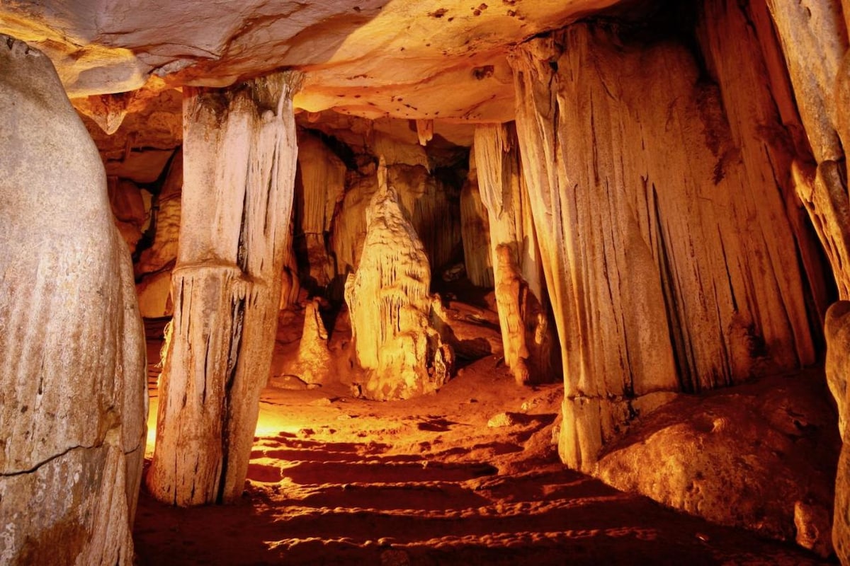 An expansive underground cave is illuminated by soft, warm lights, revealing towering rock formations and textured stone walls. Stalactites hang from the ceiling, and a series of steps leads further into the cavernous space, evoking a sense of natural wonder.