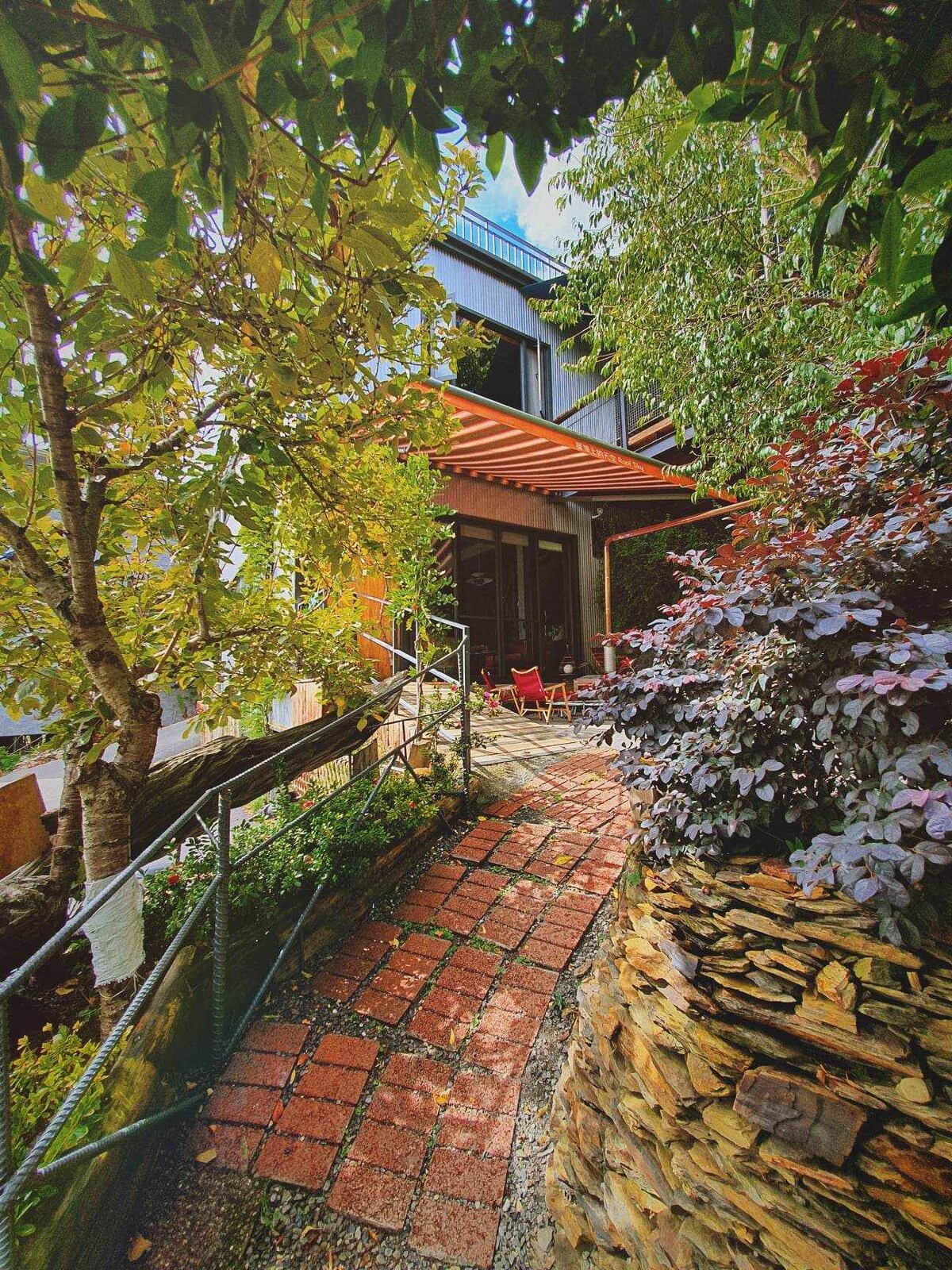 A winding brick pathway is bordered by lush greenery and colorful plants. The path leads to a modern structure featuring large glass doors, under a shaded pergola. Natural stone elements add texture to the landscape, creating a harmonious connection between the built environment and nature.