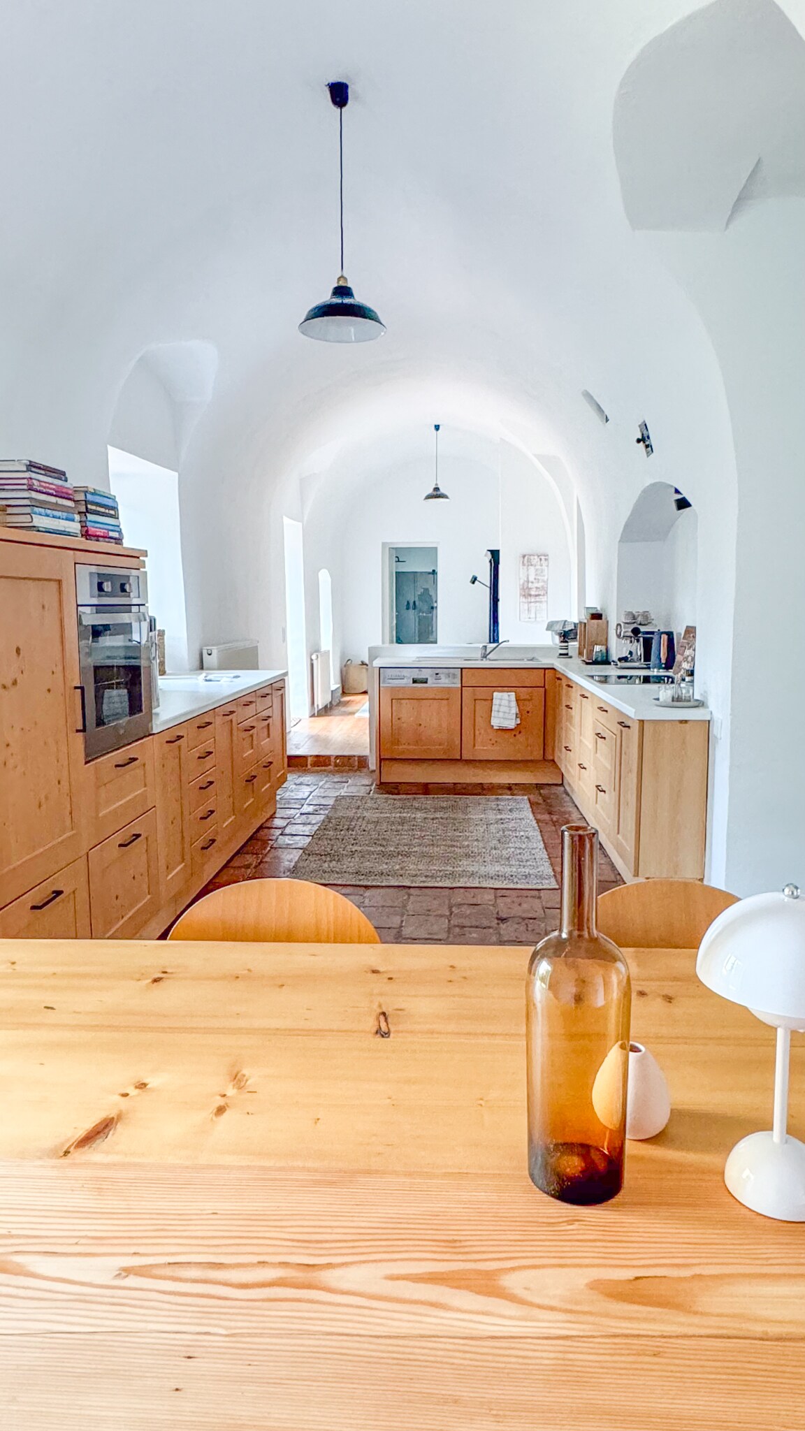 A spacious kitchen features wooden cabinetry and modern appliances, creating a functional cooking space. A wooden dining table is positioned in the foreground, accompanied by simple seating. Soft light is provided by pendant lamps hanging from the arched ceiling, enhancing the airy atmosphere.