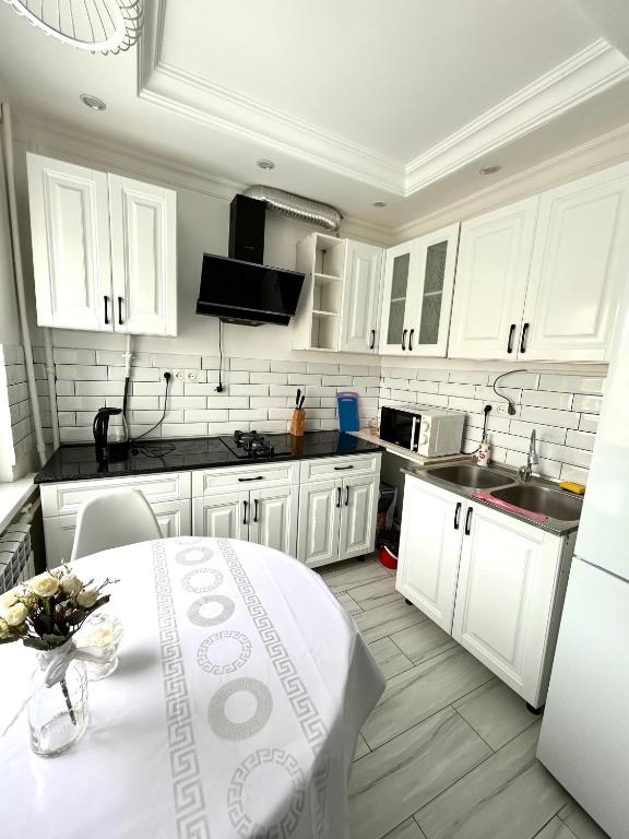 A modern kitchen features white cabinetry and a dark countertop. Appliances include a microwave, electric kettle, and a sink. A small dining table is set with a vase of flowers. Natural light enters through a window, illuminating the tiled floor and glossy surfaces.