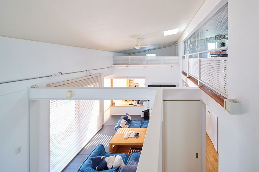 An airy living space with high ceilings is observed from an elevated perspective. A blue sofa and wooden coffee table create a comfortable seating area. Natural light streams in through the large windows adorned with sheer curtains, enhancing the open feel of the room.