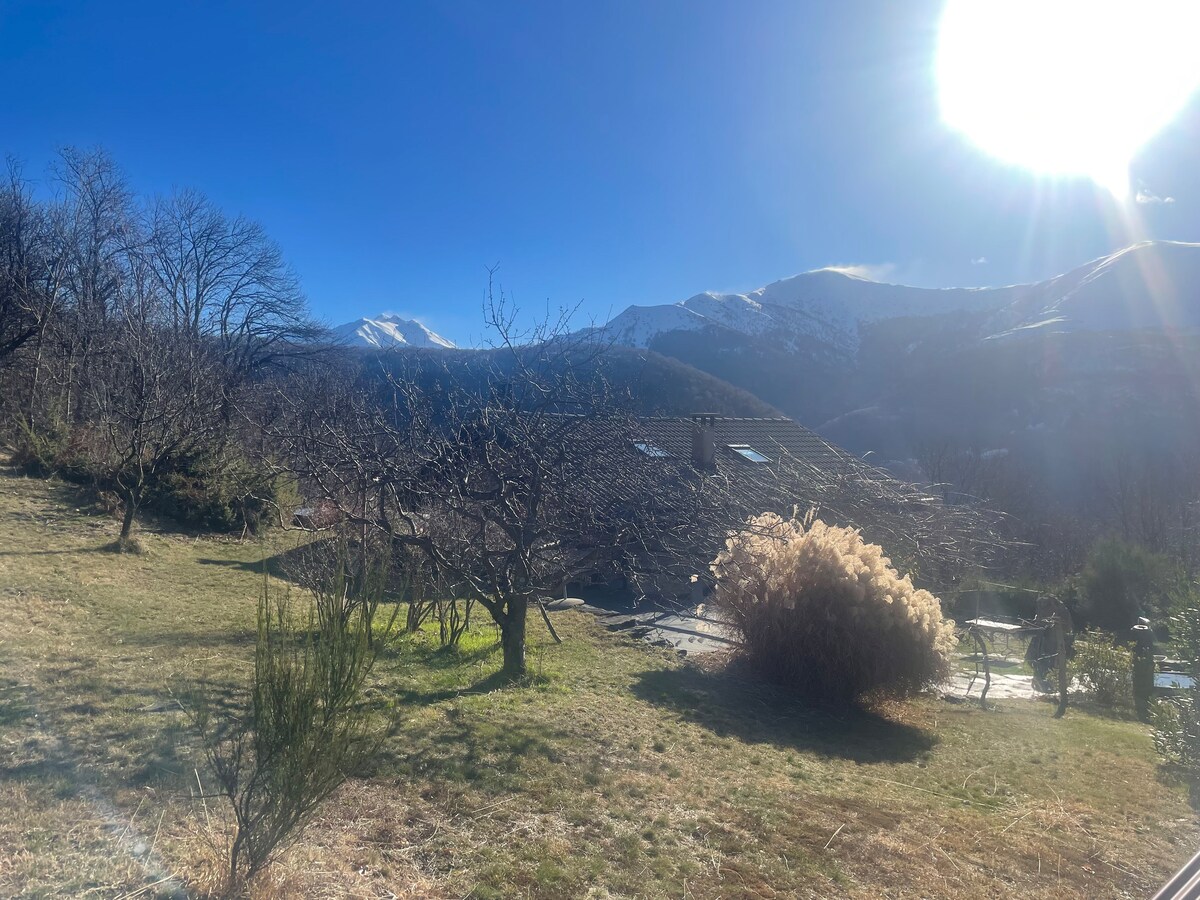 A scenic view captures a spacious outdoor area featuring a lush green landscape with trees and shrubs. The distant mountains are visible under a clear blue sky with sunlight shining brightly, creating a peaceful natural setting.