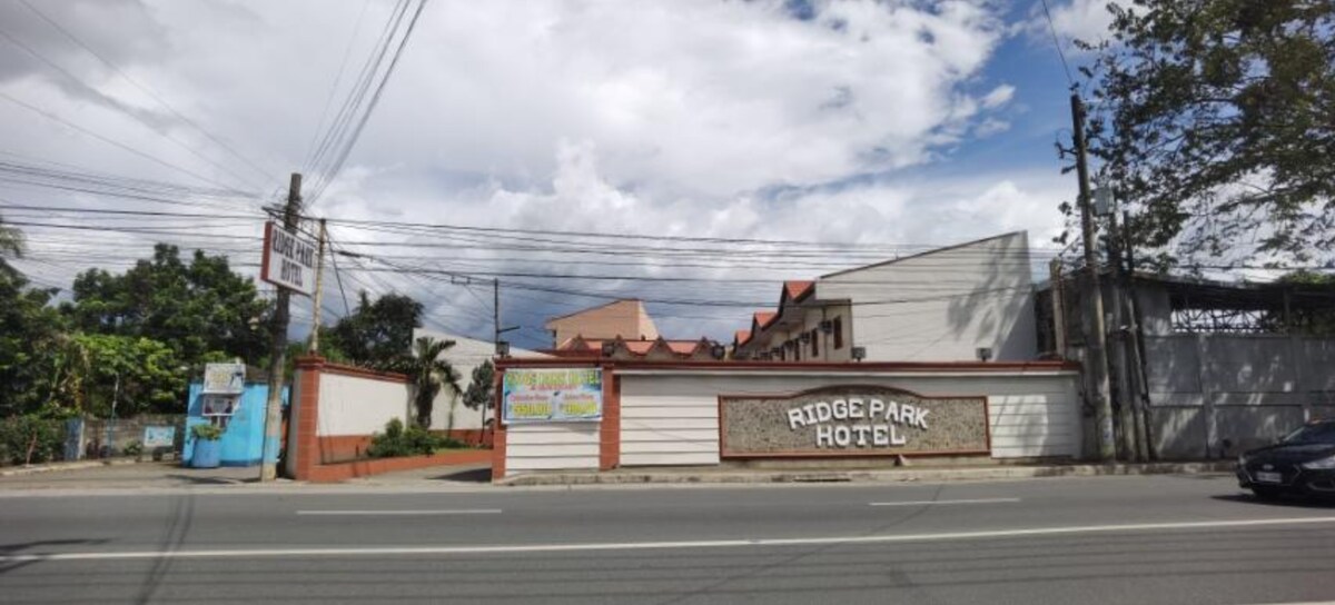 The exterior of Ridge Park Hotel is showcased, featuring a structured blend of modern and traditional architectural elements. Prominent signage displays the hotel name, bordered by a landscaped area with greenery. Power lines are visible in the background against a partly cloudy sky.
