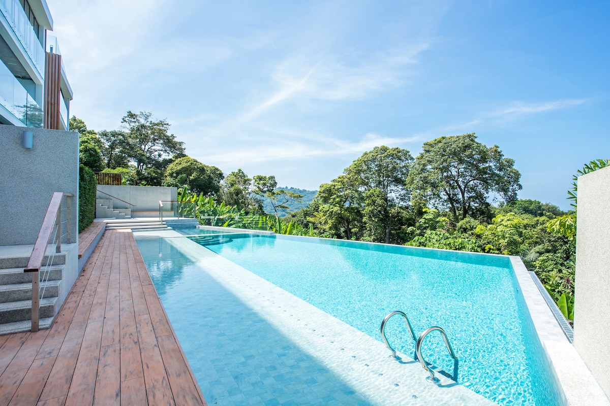 An infinity pool stretches across the image, reflecting the clear blue sky. Surrounding greenery offers a serene backdrop, enhancing the tranquil atmosphere. Steps leading down to the pool area are visible, complementing the wooden decking that borders the water.