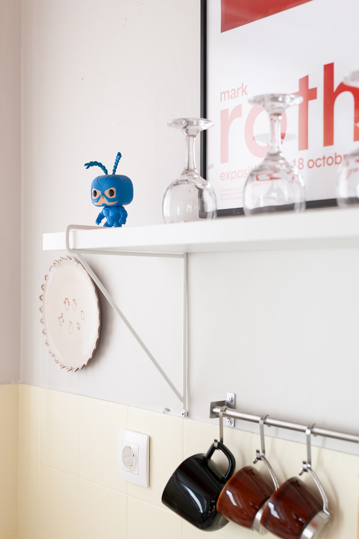 A shelf is displayed with a decorative blue figure beside neatly arranged glassware. A collection of plates and cups hangs below, showcasing a variety of designs. The simple wall structure is complemented by modern lighting, emphasizing the clean lines of the space.