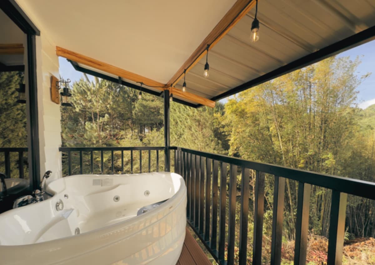 A private Jacuzzi is located on a wooden deck, surrounded by lush greenery. Soft lighting is provided by hanging bulbs above, creating a serene ambiance. The balcony railing frames a scenic view of the trees and natural landscape beyond.