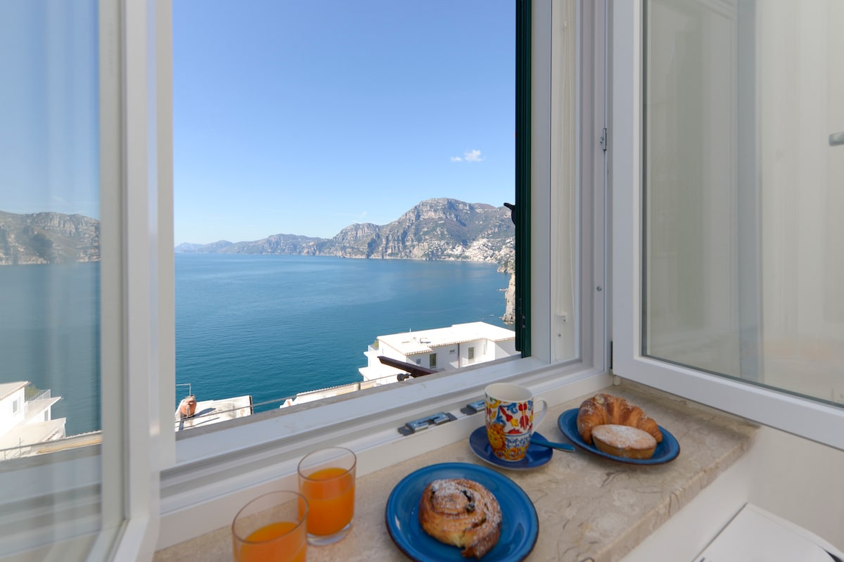 A view from the window showcases the sparkling blue sea and hilly coastline. A tray with pastries and drinks rests on the sill, contributing to the serene atmosphere. Natural light floods the space, enhancing the scenic backdrop.