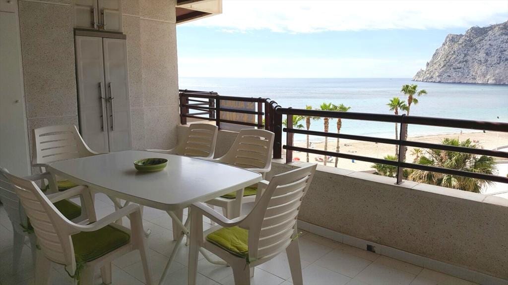 A spacious terrace features a large dining table surrounded by six white chairs with green cushions. The view includes sandy beach and palm trees, framed by distant cliffs under a partly cloudy sky.