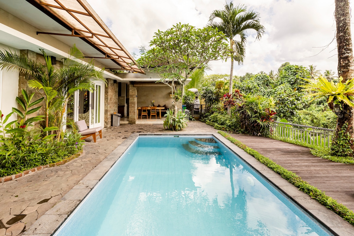 A private pool is showcased, surrounded by tropical greenery and plants. A stone pathway leads to the pool area, with a seating arrangement visible nearby. The villa's exterior features large windows and a covered patio, providing a seamless connection to the lush landscape.