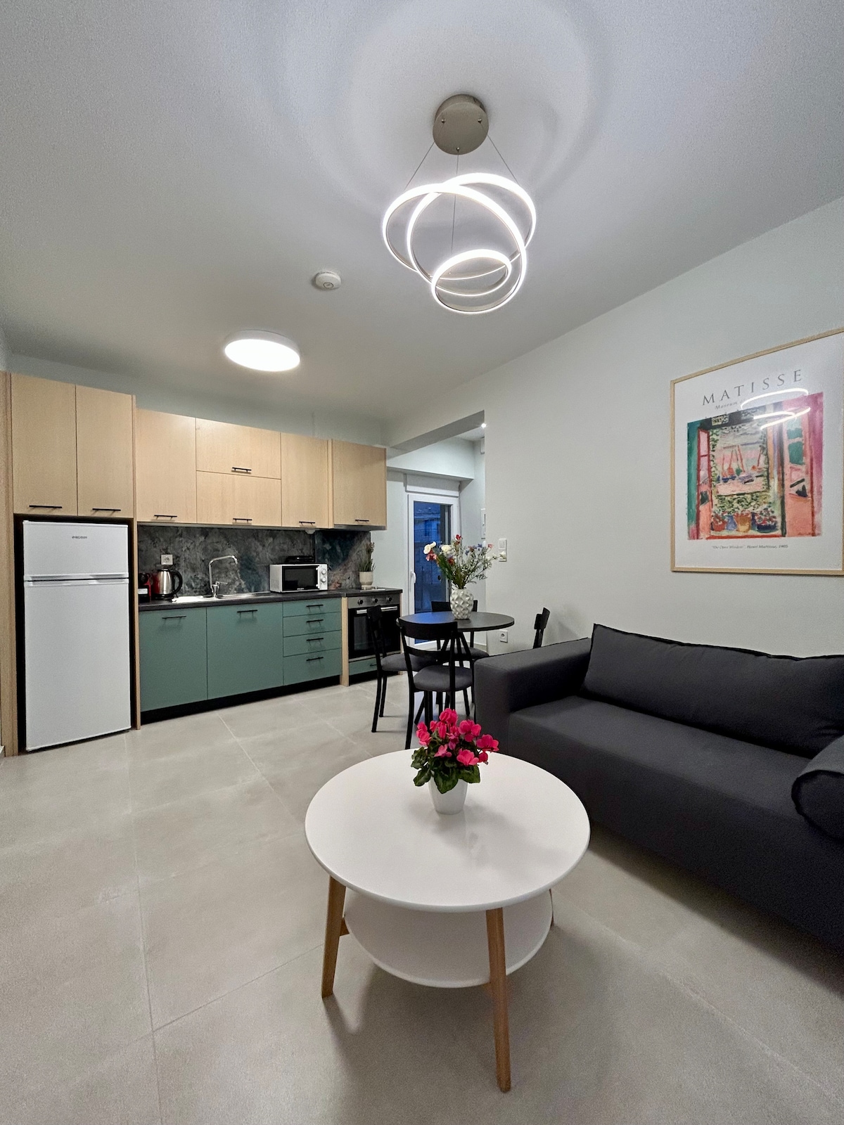 A modern kitchen and living area feature a black sofa and a round coffee table adorned with pink flowers. Storage and appliances in the kitchenette are complemented by light wood cabinetry and dark granite countertops, creating a cozy yet functional space.