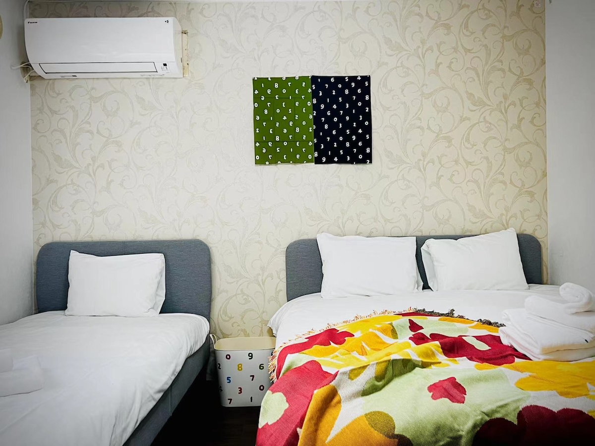 The room features two beds, one spacious double bed with a vibrant blanket and a single bed. A neutral patterned wall serves as a backdrop, while a pair of colorful wall art pieces adds visual interest. A small decorative element is visible near the floor.