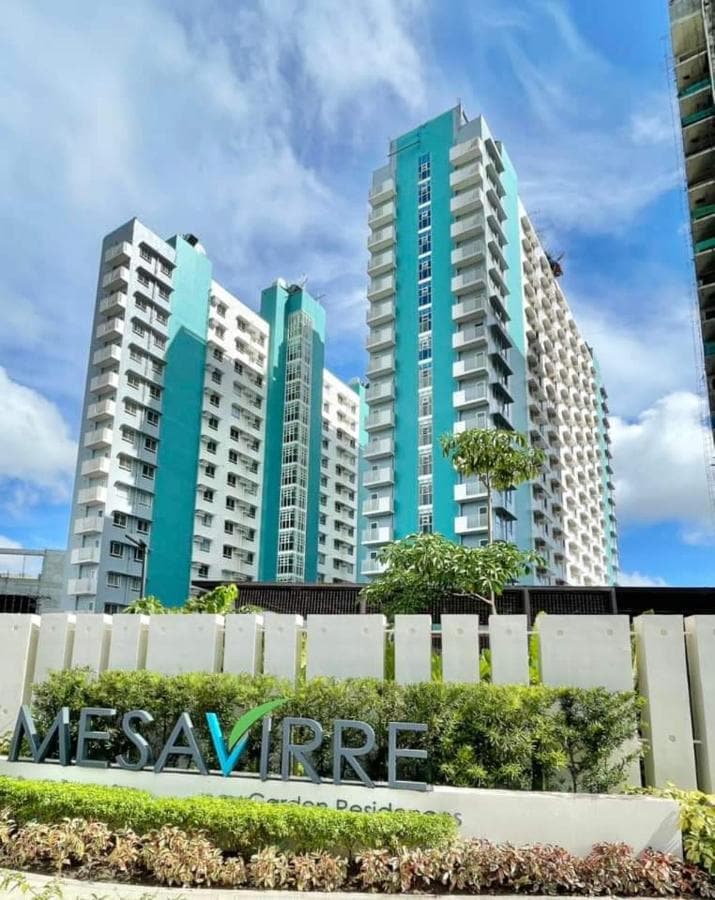 The exterior of Mesavirre Garden Residences is displayed, featuring modern high-rise buildings with a vibrant turquoise accent. Lush greenery surrounds the property, and a sign with the name 'MESAVIRRE' is prominently positioned at the front, complementing the contemporary architecture.