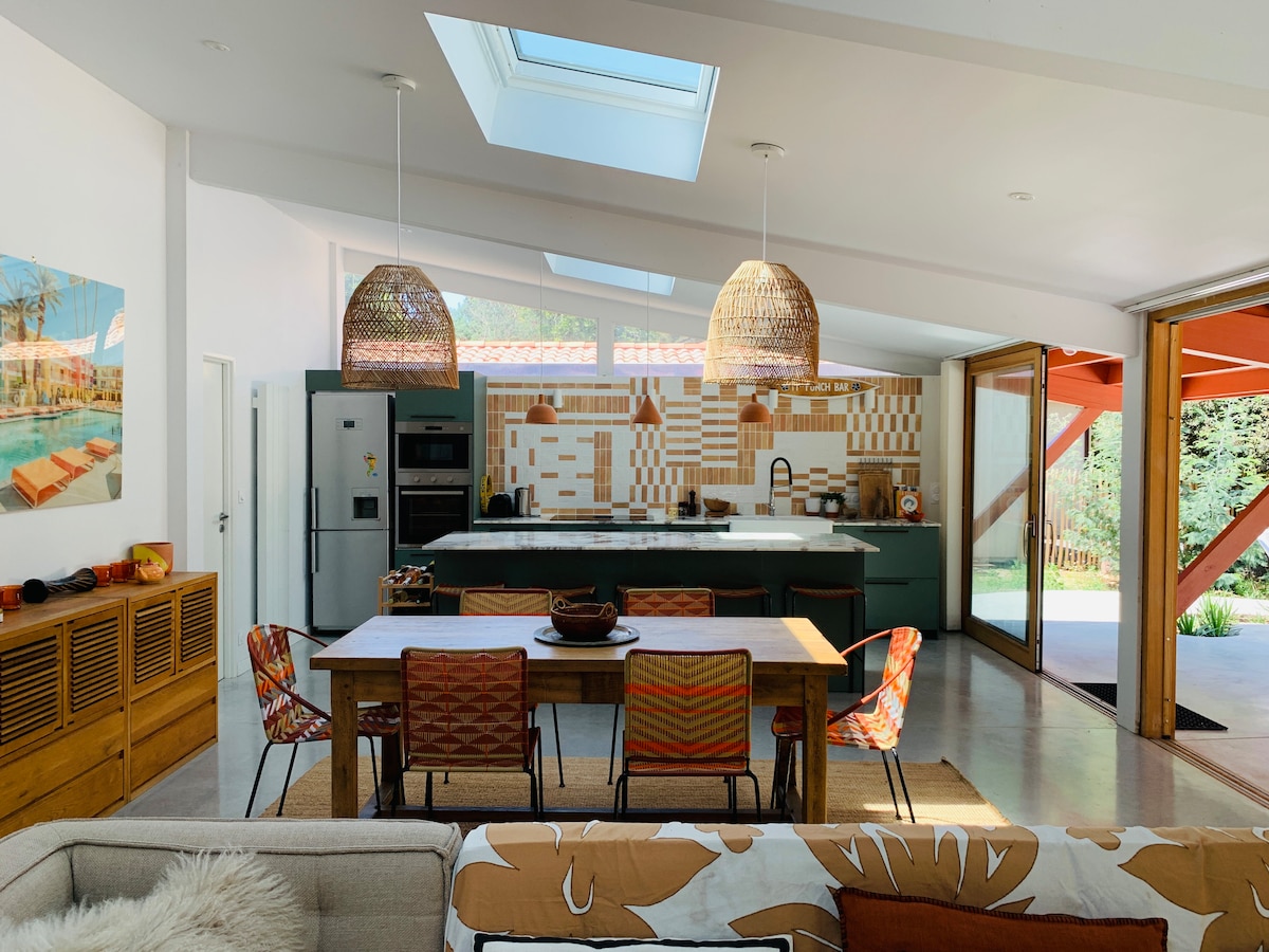 An airy kitchen is showcased with a high ceiling and skylights, allowing natural light to fill the space. Modern appliances and patterned backsplash are present. A spacious dining table is surrounded by colorful chairs, and large glass doors open to the outdoor area.