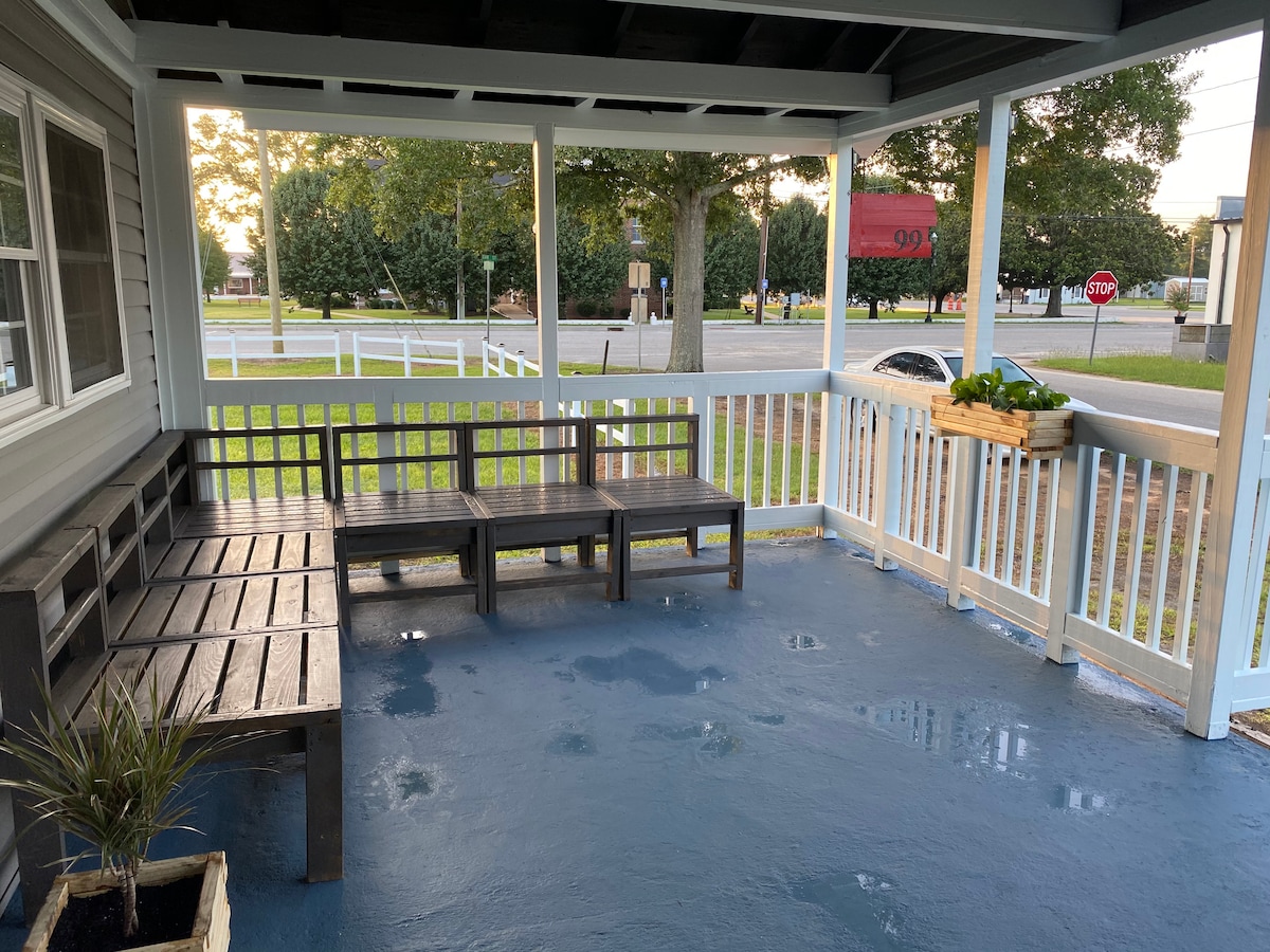 A spacious porch features rustic wooden seating arranged for relaxation. A potted plant adds a touch of greenery. The sturdy railings provide safety, while the surrounding view includes greenery and a nearby road, ensuring a pleasant outdoor experience.