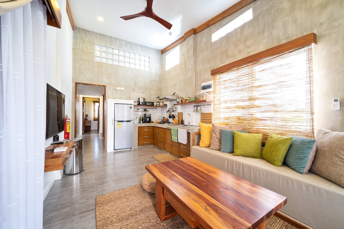 A spacious living area features a comfortable seating arrangement with variously colored cushions, a large wooden coffee table, and an adjacent kitchen equipped with modern amenities. Natural light floods the space through a wide window adorned with bamboo blinds, creating a relaxed, welcoming environment.