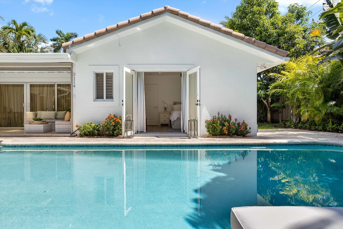 A charming exterior of the beach house is highlighted by glass double doors that open to a serene indoor space. A tranquil pool reflects the clear blue sky, surrounded by lush greenery and flowering plants, creating a relaxed outdoor atmosphere.