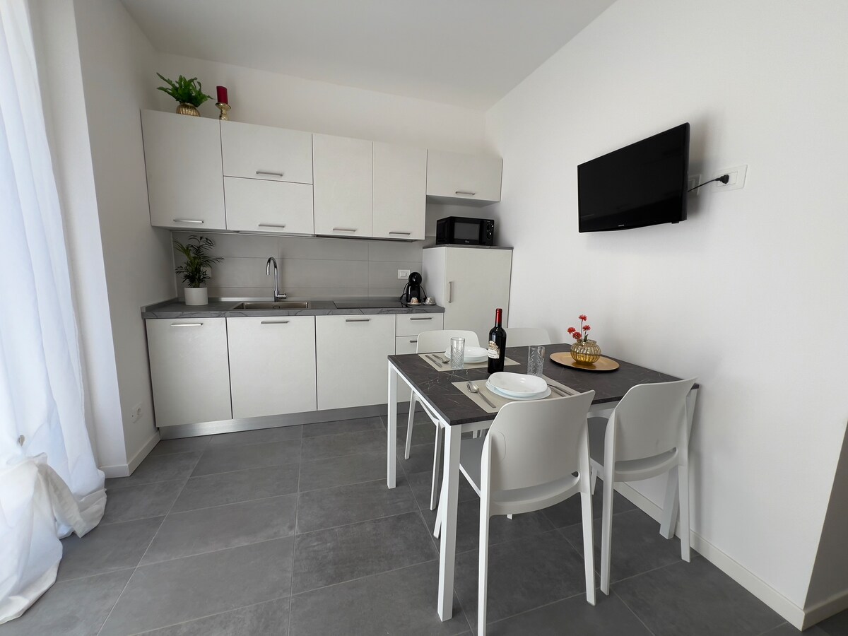 The kitchen area features modern cabinetry and appliances, including a microwave and coffee maker. A dining table is set with tableware on a gray surface, accompanied by two white chairs. Natural light fills the space through a large window, enhancing the clean and functional design.