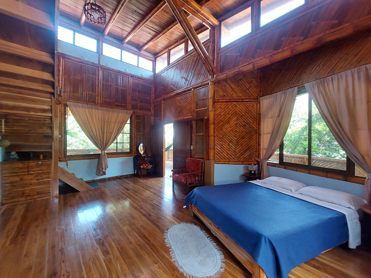 A spacious room features high wooden ceilings and large windows that allow natural light to filter in. A cozy bed is dressed in a blue coverlet, with soft white linens. A comfortable armchair sits nearby, with a stylish mirror reflecting the room’s rustic charm.