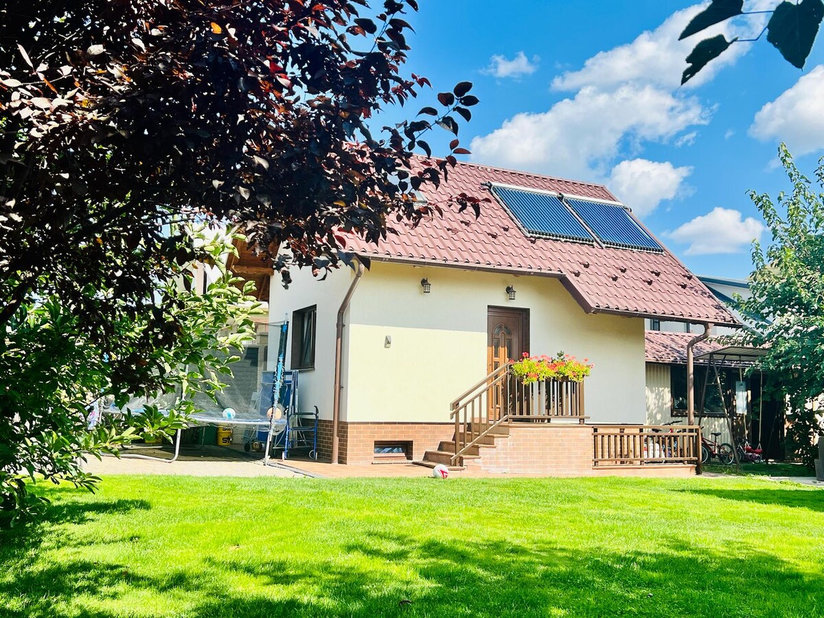 A charming house with a sloped roof, featuring solar panels and surrounded by lush greenery. The entrance is highlighted by a small staircase and flower pots. A spacious lawn extends in front, with trees providing gentle shade and a bright blue sky overhead.