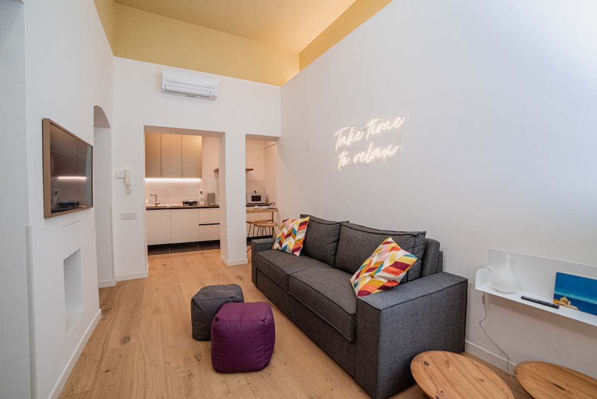 The living area features a spacious gray sofa adorned with colorful cushions. A wall-mounted television is visible, alongside a compact dining area that leads to a fully equipped kitchen. Soft lighting enhances the relaxing ambiance, complemented by a small table and decorative poufs.