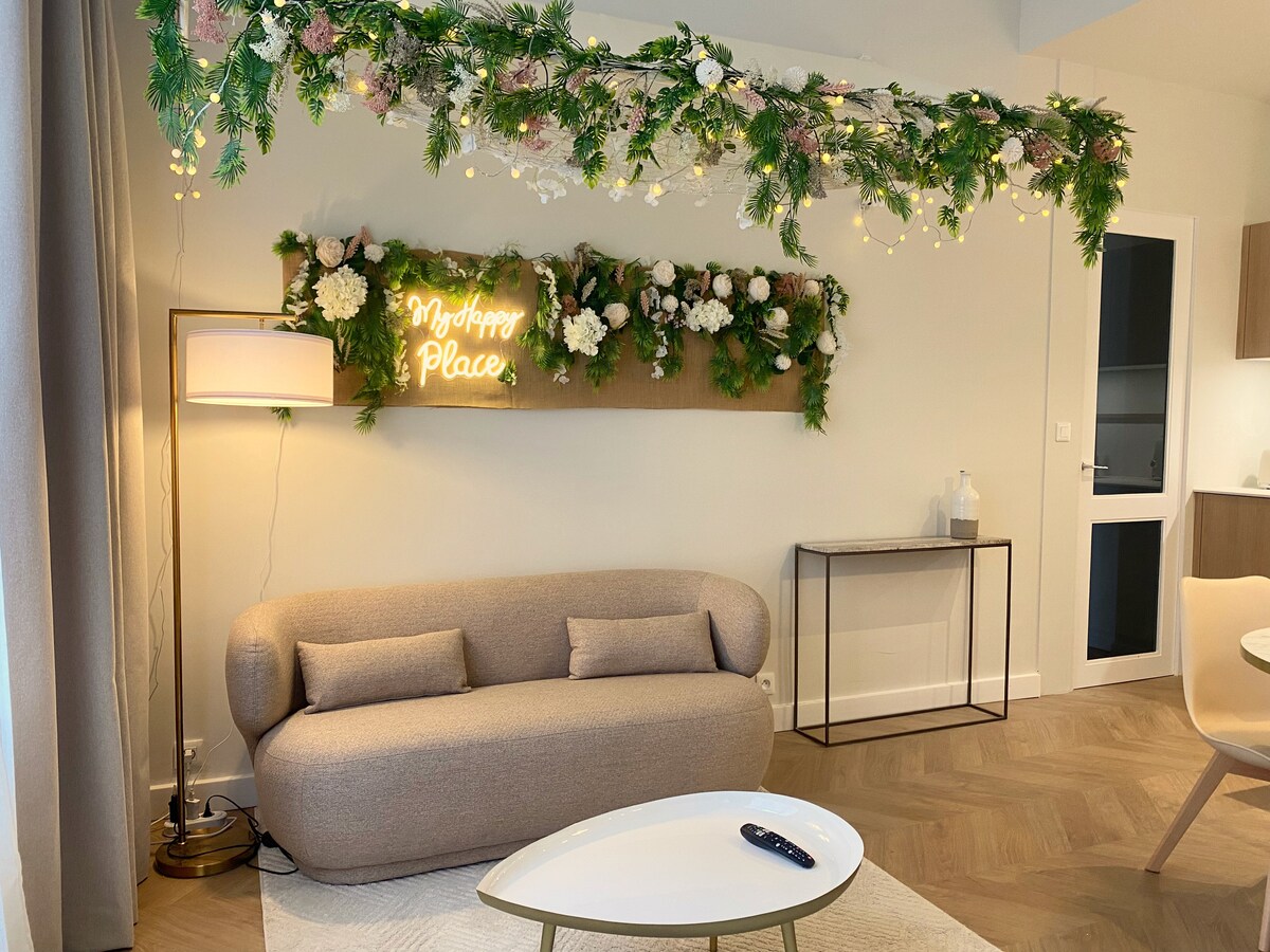A softly curved sofa is positioned across from a stylish coffee table, featuring a remote control placed on its surface. Decorative greenery and fairy lights adorn the wall above, with a neon sign reading 'My Happy Place.' A lamp provides warm lighting nearby.