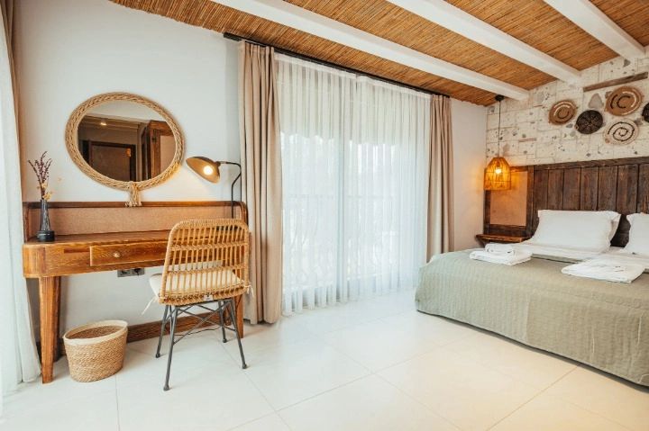 A spacious room features a cozy bed with neutral bedding and two pillows. A wooden desk and chair provide a work station, illuminated by a stylish lamp. Floor-to-ceiling windows are dressed with sheer curtains, allowing natural light to fill the space.