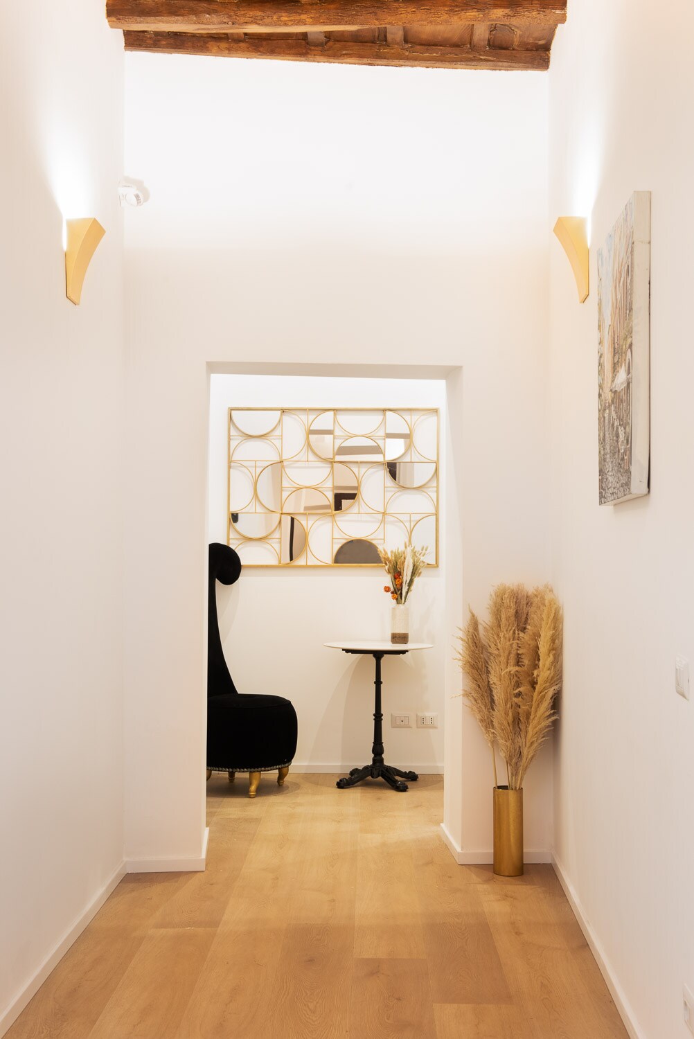 A well-lit hallway features white walls and warm wooden flooring. A decorative wall mirror with geometric designs reflects light. A small table, paired with a stylish black chair, is positioned beside a tall, decorative plant, adding a touch of elegance to the space.
