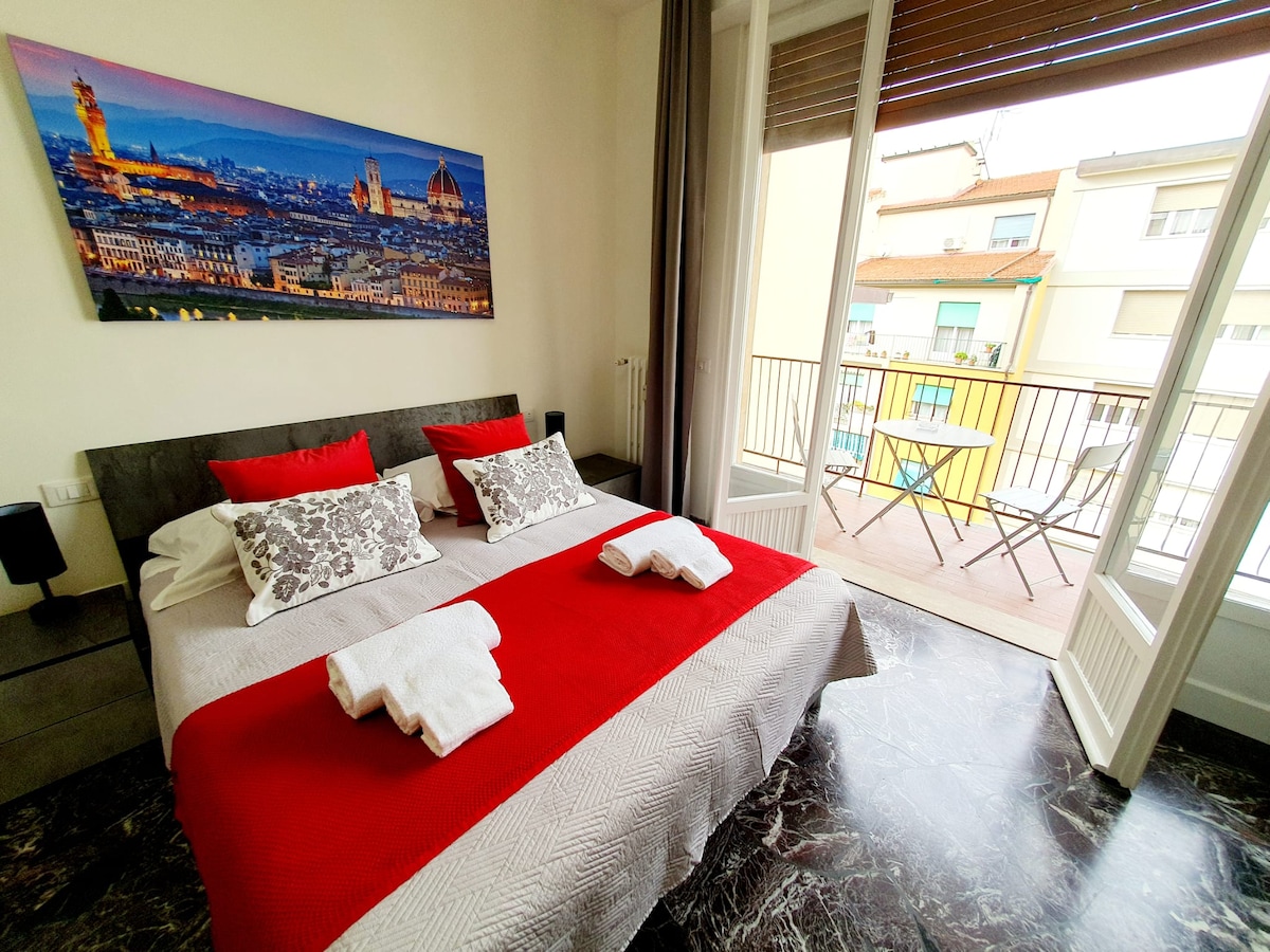 A spacious bedroom features a large bed adorned with grey and white linens, complemented by decorative pillows in red. A vibrant cityscape artwork captures the charm of Florence above the bed. Natural light floods in through a wide balcony door, which opens to a small outdoor table and chairs.