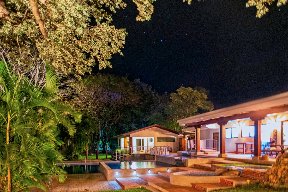 The night scene reveals a serene farmhouse surrounded by lush greenery. A well-lit private pool reflects the moonlight, while an outdoor dining area is visible beside the house. Stars twinkle overhead, enhancing the tranquil atmosphere of the expansive property.