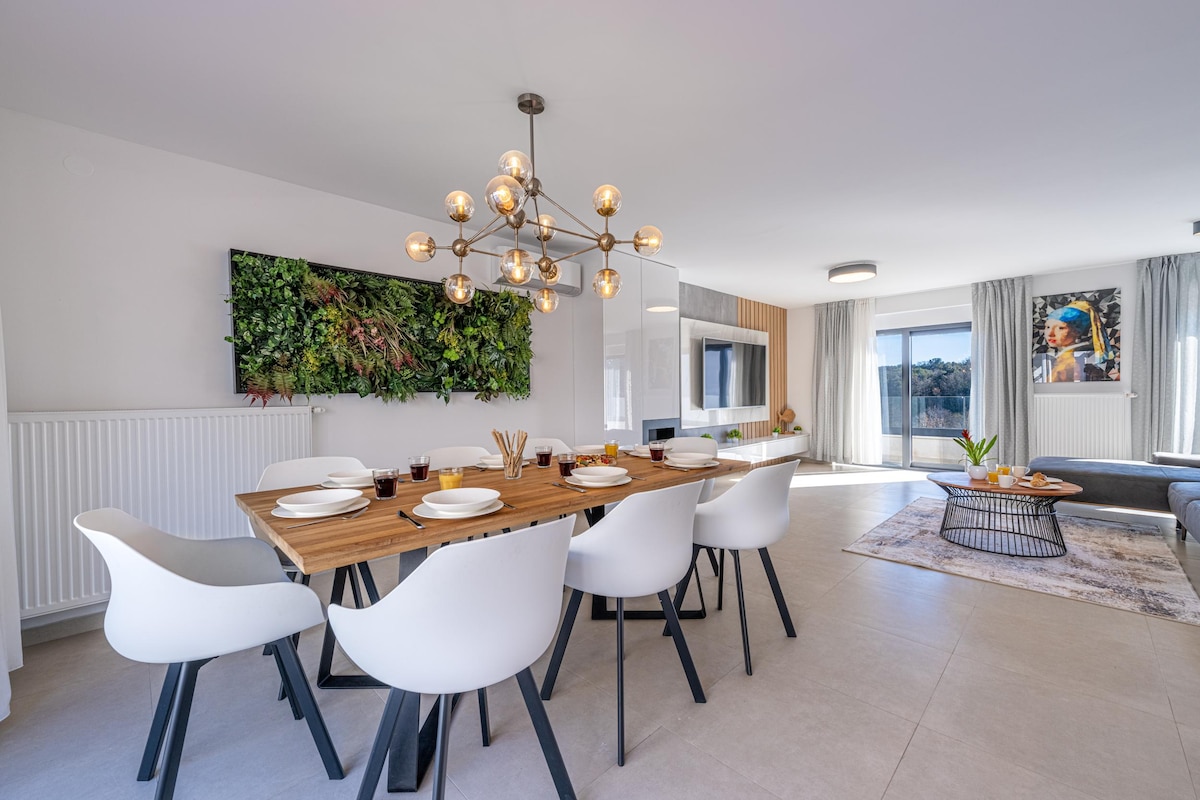 A modern dining area features a wooden table surrounded by six white chairs. A unique light fixture hangs overhead, and a vertical garden is displayed on the wall. Natural light filters through large windows, illuminating the adjacent living space with soft gray accents.