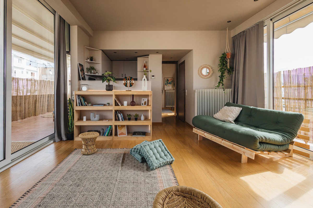 A spacious living area is highlighted by large glass doors that lead to a balcony. A green sofa is positioned against a wooden bookshelf filled with plants and books. Natural light fills the room, which features warm wood flooring and neutral tones in the decor.