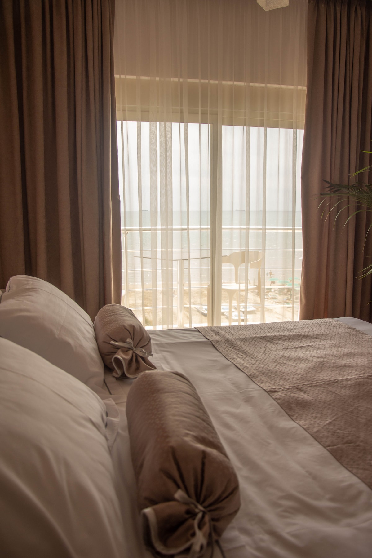 A serene bedroom features a spacious bed dressed with crisp white linens and decorative bolster pillows. Soft, flowing curtains frame the sliding glass doors that lead to a balcony. Ocean views are visible through the sheer fabric, with hints of outdoor furniture seen beyond.