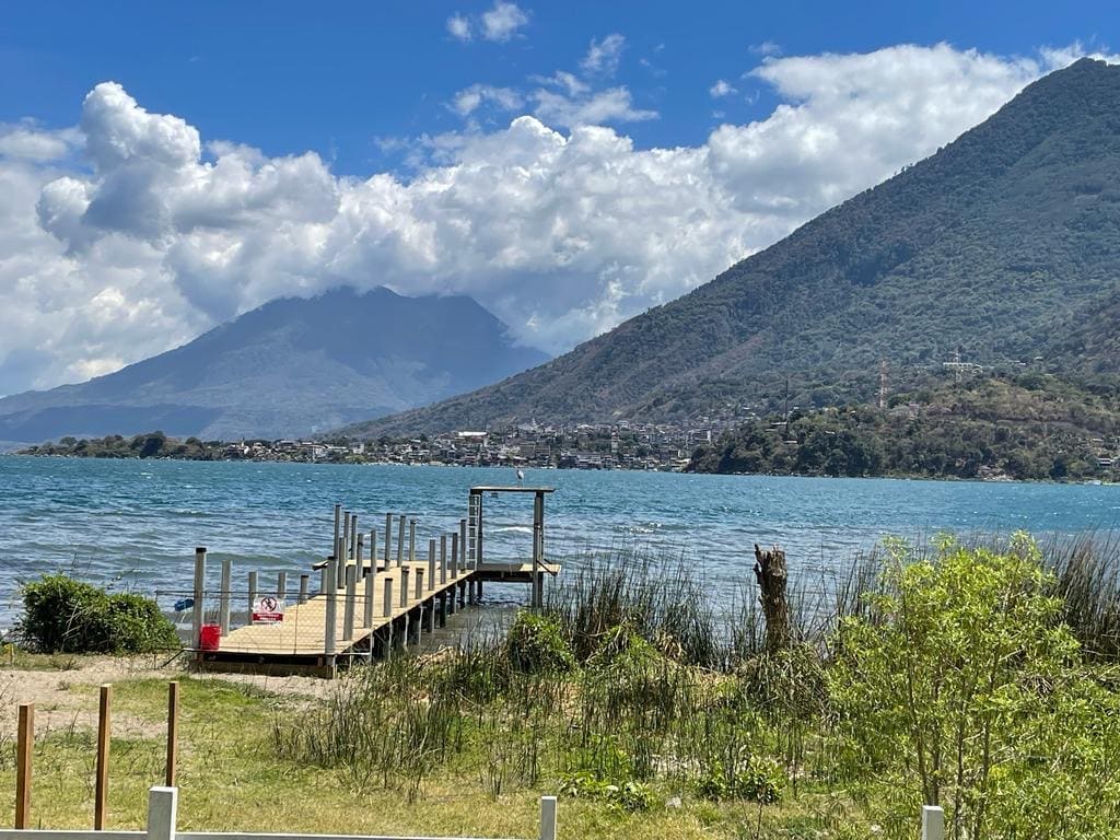 A wooden dock extends into the vibrant blue waters of Lake Atitlan, surrounded by lush greenery and mountains. Fluffy clouds drift above, casting gentle shadows on the landscape. The tranquil scene invites opportunities for water activities and relaxation in a serene natural setting.